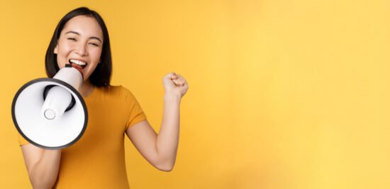Smiling asian woman standing with megaphone, announcing smth, advertising product, standing over yellow background